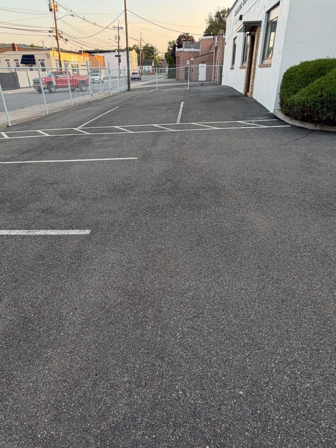 Commercial parking area before surface work beside a white building and chain-link fence.